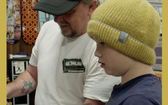 Image has a gold border and is of Rod O'Hara owner of Bellingen Video Connection in his store showing a young customer a collectible in the documentary Return Chute: The survival of a small town video store