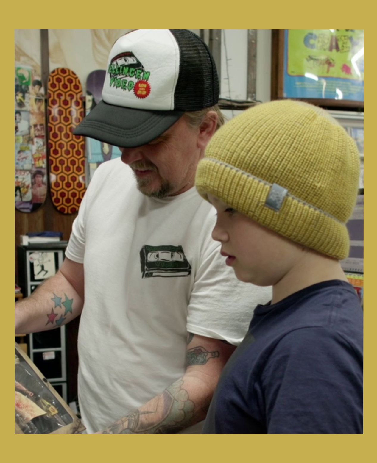 Image has a gold border and is of Rod O'Hara owner of Bellingen Video Connection in his store showing a young customer a collectible in the documentary Return Chute: The survival of a small town video store