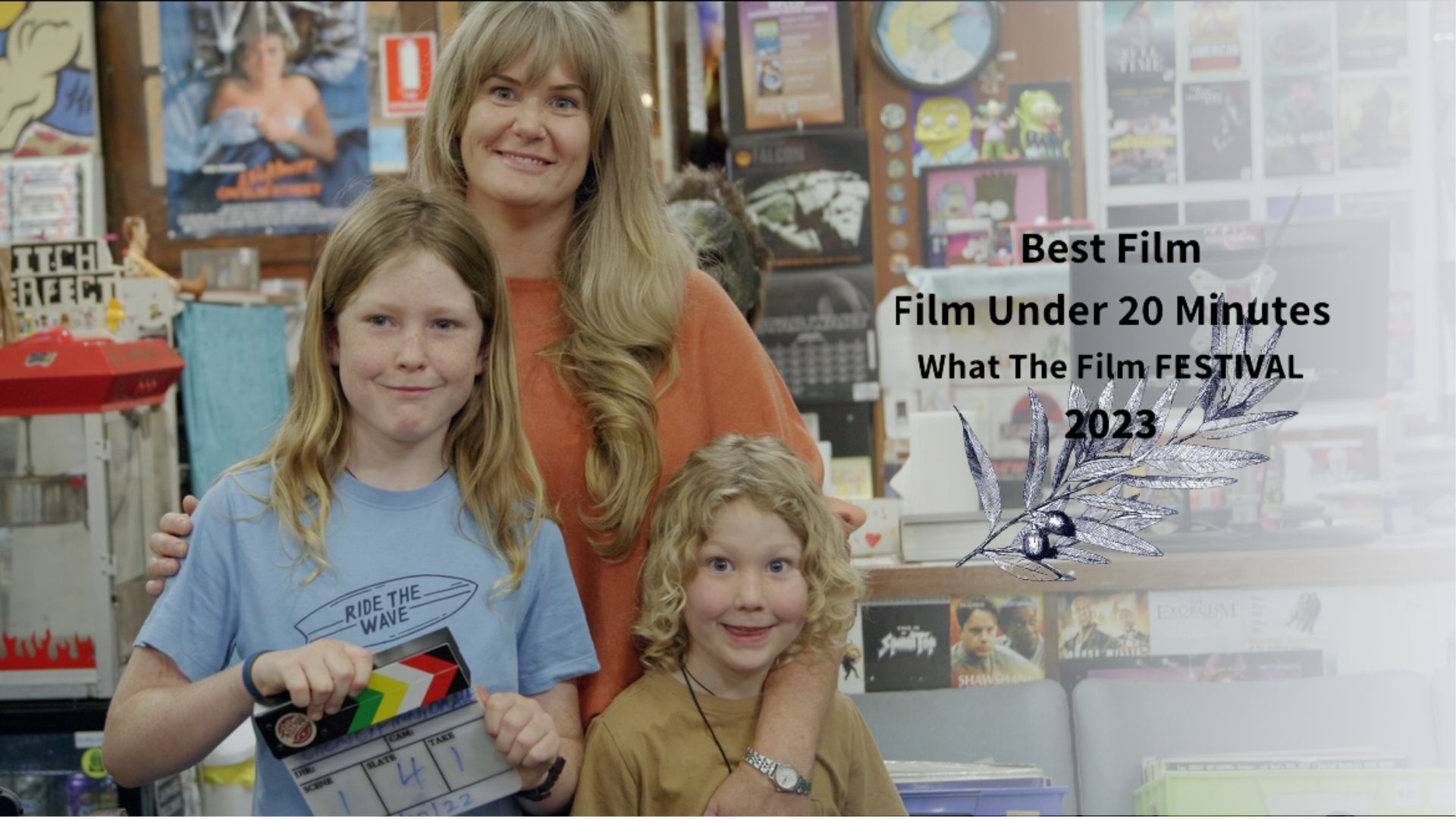Image has a gold border and woman stands with her two young children, one holding a film clapper board in a video store from the documentary Return Chute: The survival of a small town video store with a winning laurel and text in image "Best Film Under 20 minutes What The Film Festival 2023"