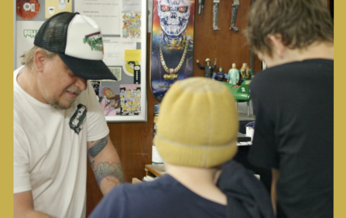 Image has a gold border and is of Rod O'Hara owner of Bellingen Video Connection in his store with two young customers facing away from the camera from the documentary Return Chute: The survival of a small town video store