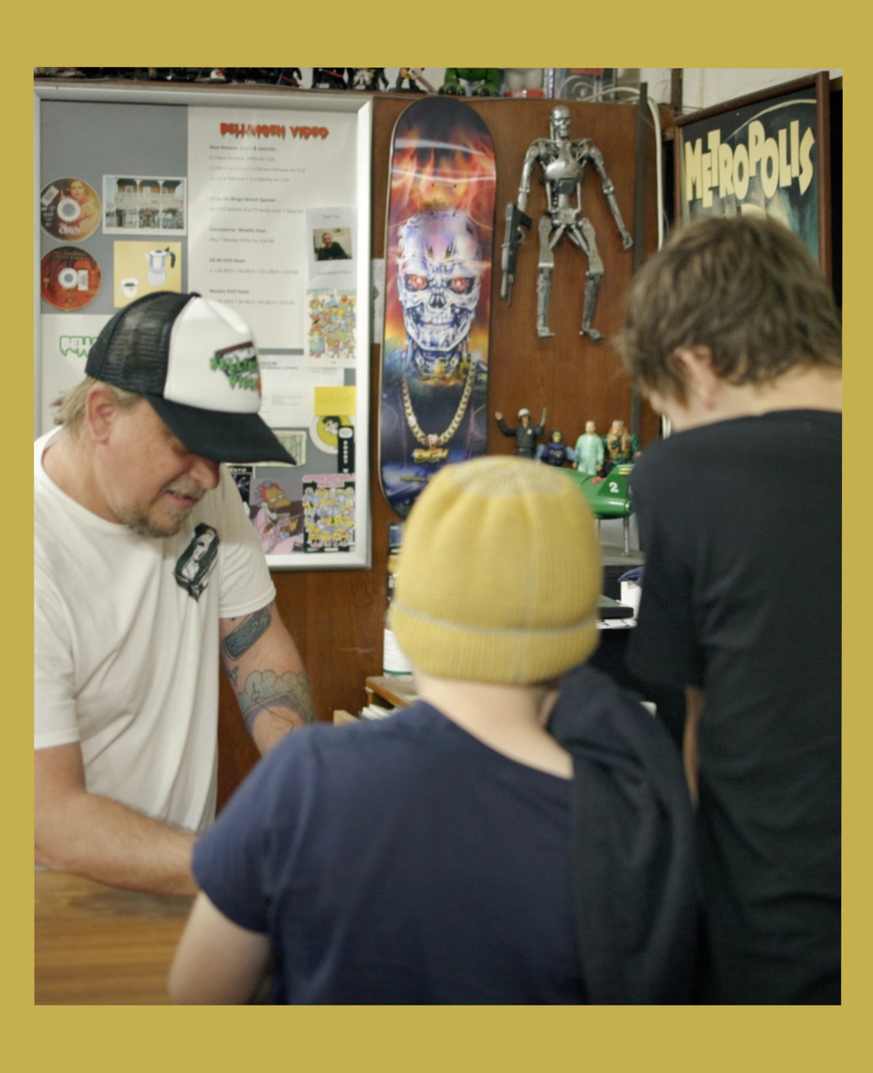 Image has a gold border and is of Rod O'Hara owner of Bellingen Video Connection in his store with two young customers facing away from the camera from the documentary Return Chute: The survival of a small town video store