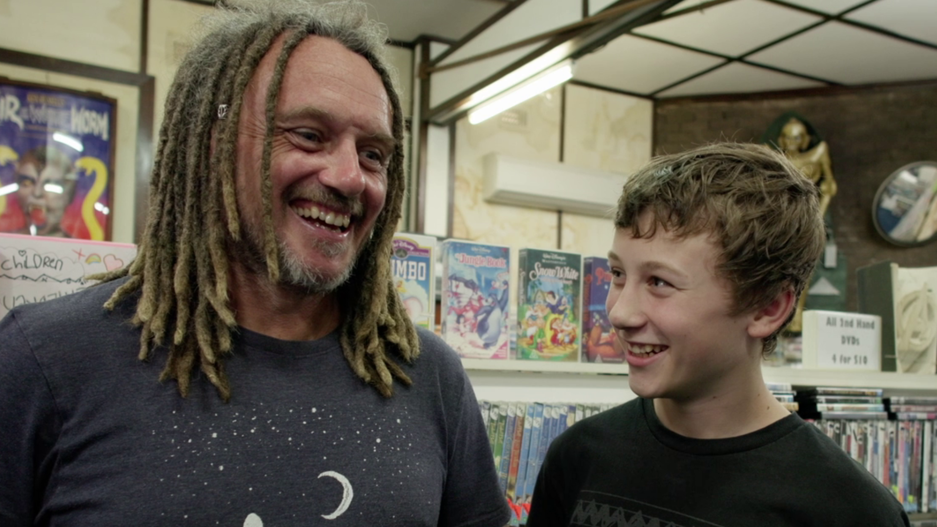 A man and his son laugh together in front of shelves of videos laughing from the documentary Return Chute: The survival of a small town video store.