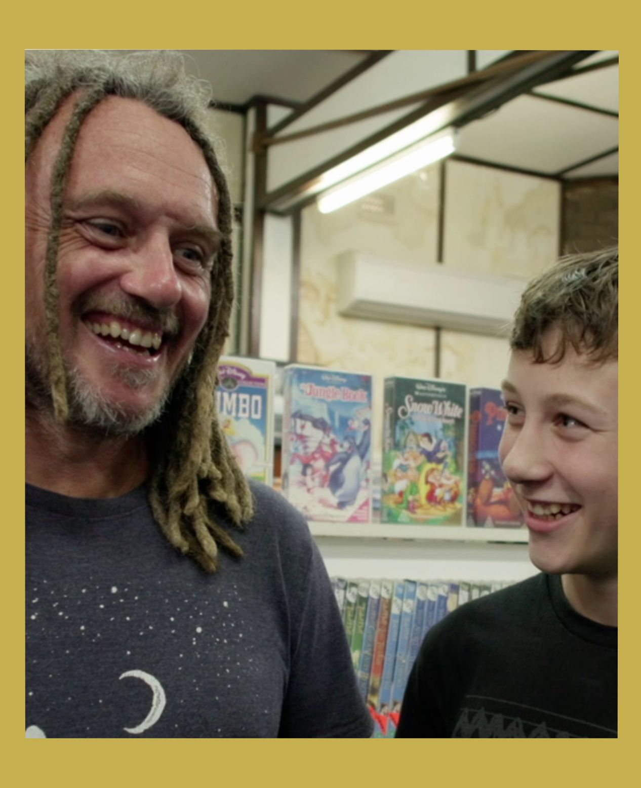 Image has a gold border A man and his son laugh together in front of shelves of videos laughing from the documentary Return Chute: The survival of a small town video store.
