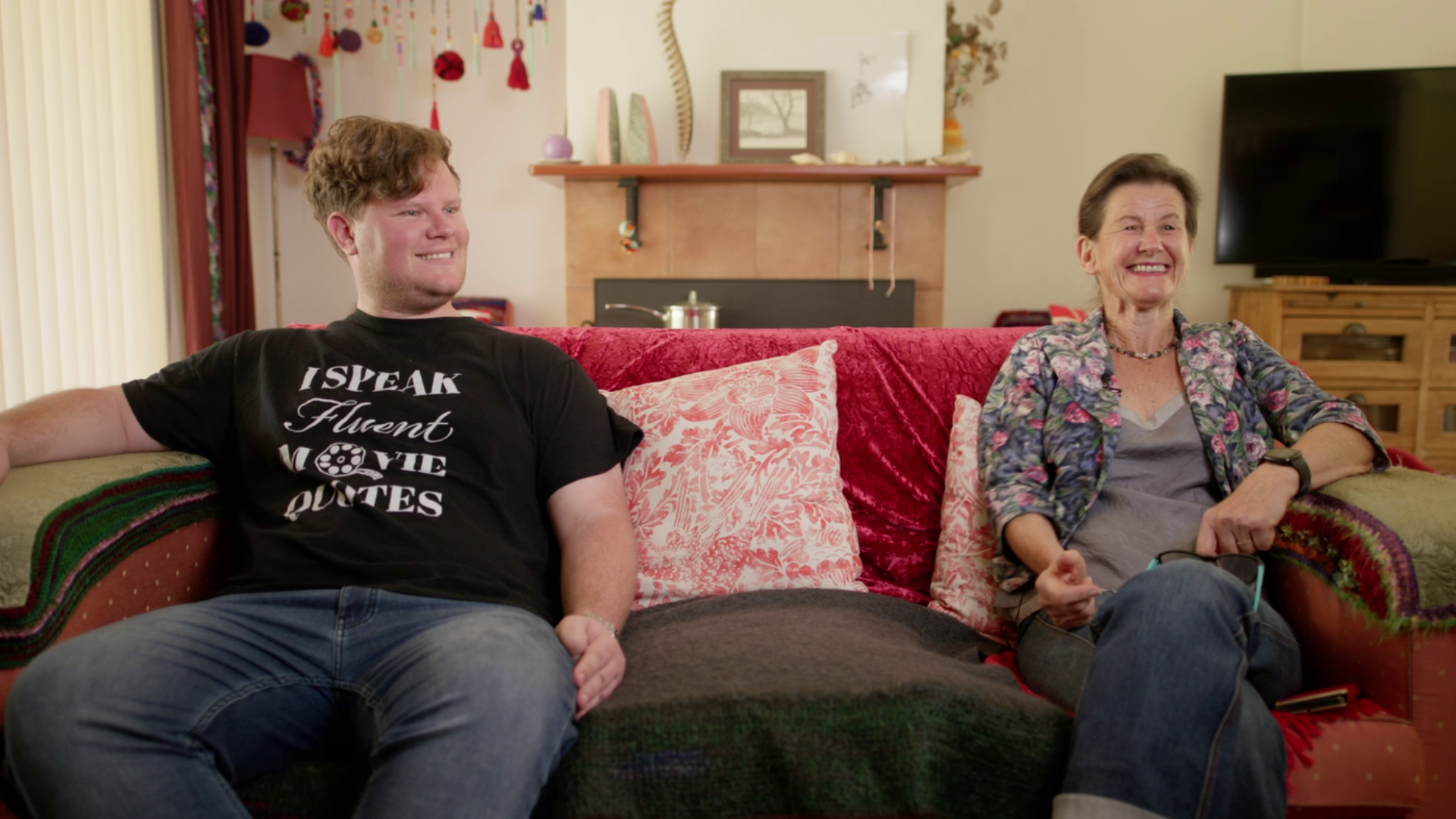 A young man and his mother, an older woman sit on a couch in their home laughing. The young man wears a shirt that says: I speak fluent movie quotes.