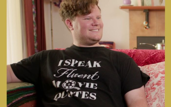 Image has a gold border. A young man sits on a couch smiling to someone out of frame. He wears a shirt that says: I speak fluent movie quotes.
