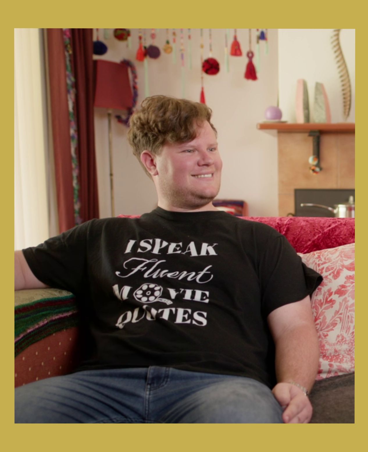 Image has a gold border. A young man sits on a couch smiling to someone out of frame. He wears a shirt that says: I speak fluent movie quotes.