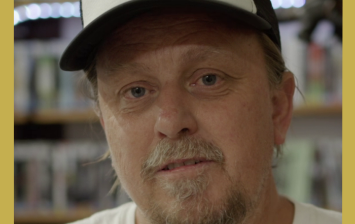 Image has a gold border and is of Rod O'Hara owner of Bellingen Video Connection in his store looking at camera in the documentary Return Chute: The survival of a small town video store