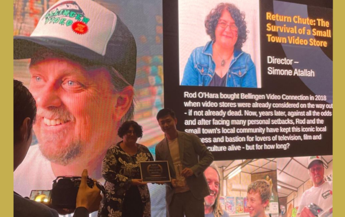 Documentary Return Chute: The Survival of a small town video store director and producer Simone Atallah stands in front of a large screen to receive the Special Jury Award at the Sydfest Independent FIlm Festival 2024. The image has a god border.